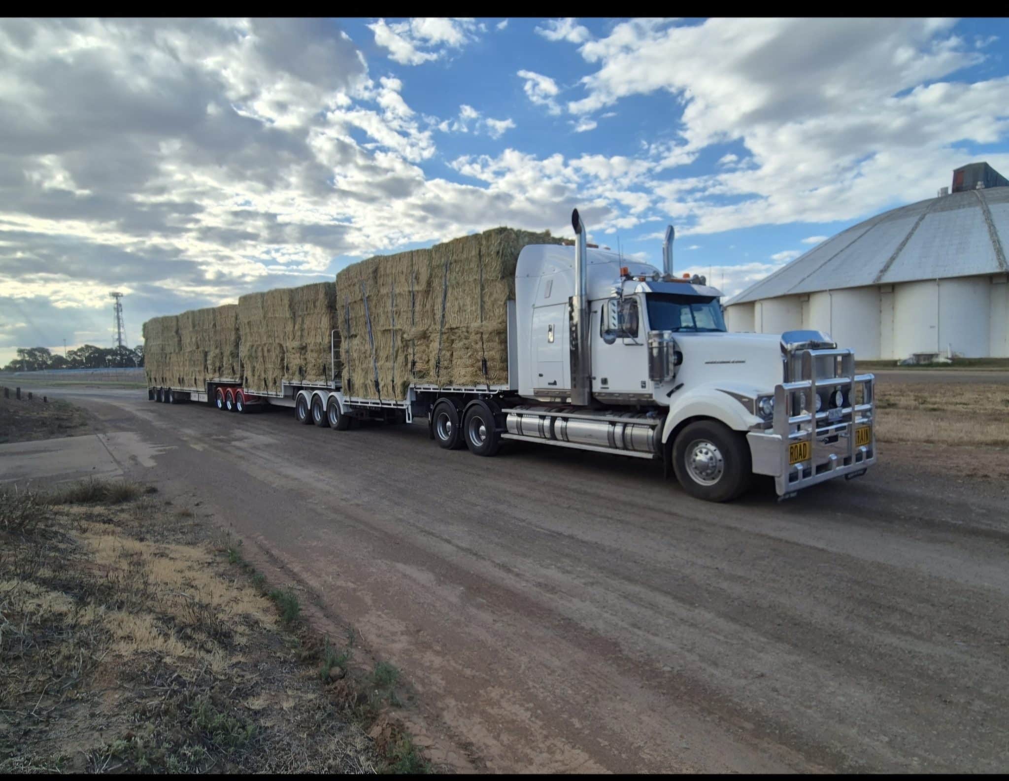 White semi-truck hauling a long flatbed loaded with stacked hay bales beside a barn under a cloudy blue sky.