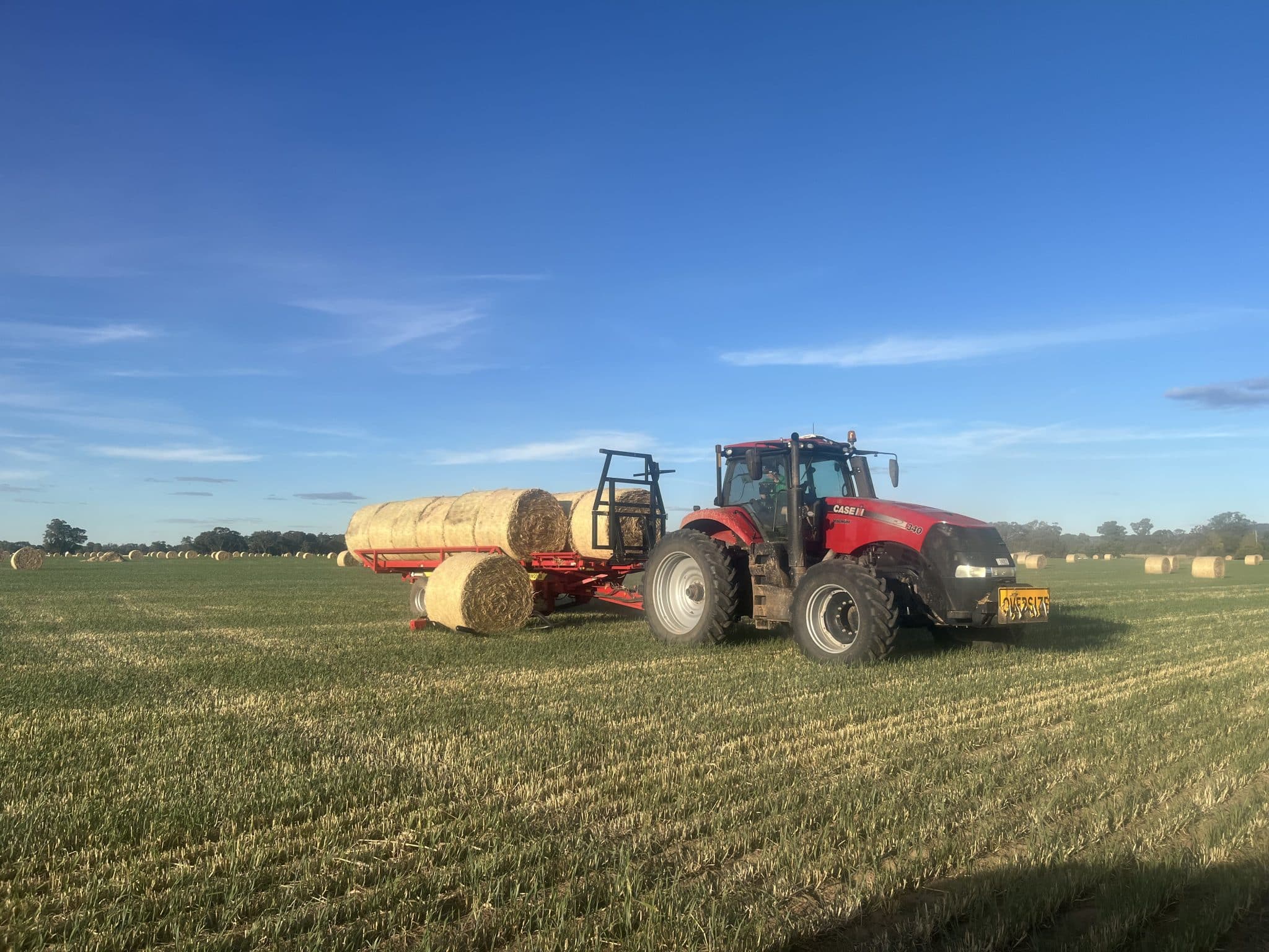 Red tractor pulling a hay bale trailer in a wide field, with round hay bales and clear blue sky above.