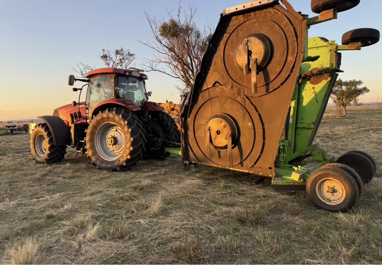 Red tractor with large front tires attached to a green round-bale baler in a dry field at sunset, large equipment resting on the grass.