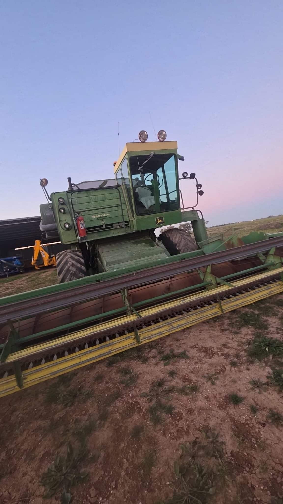 Green John Deere combine harvester with header attached, parked on a dirt field at dusk.