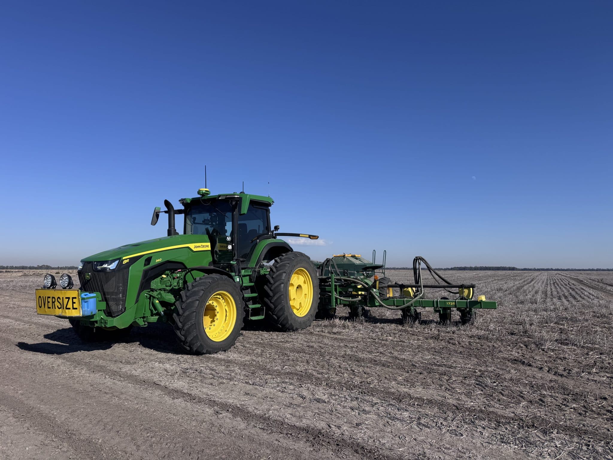 Green John Deere tractor with oversized yellow wheels parked in a plowed field under a clear blue sky, with equipment attached behind it.