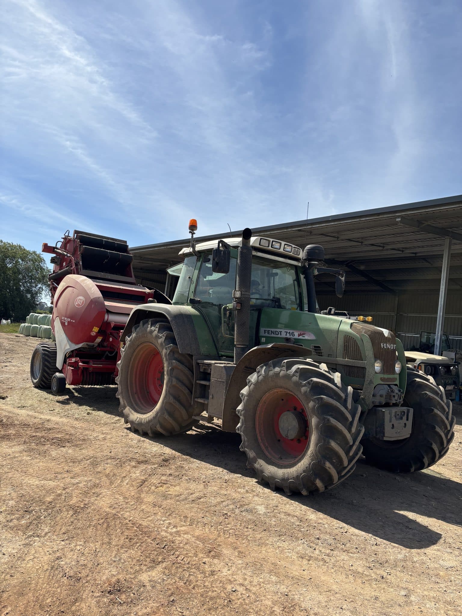 Green tractor with large tires parked beside a red baler under a metal shed, on a dirt lot with clear blue sky behind.
