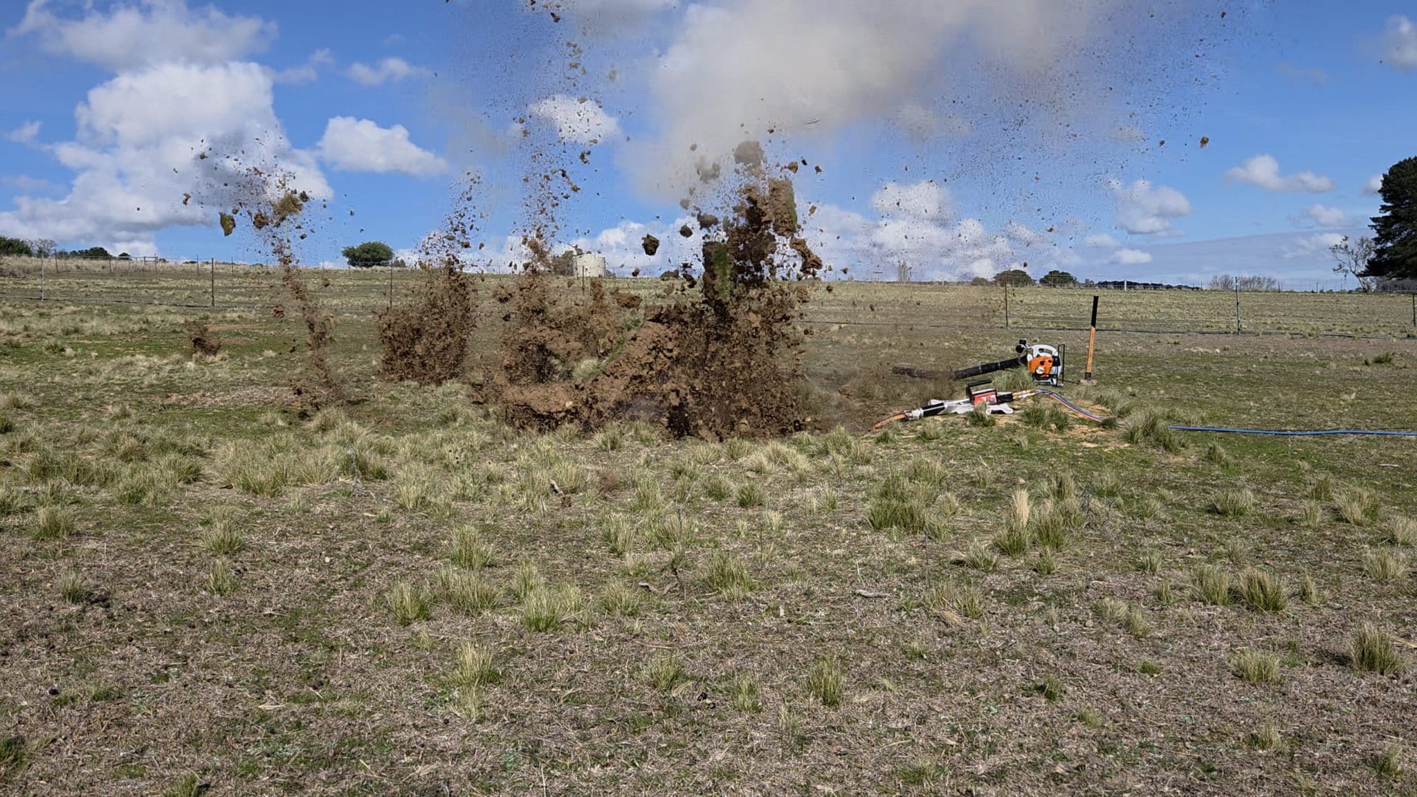 Soil eruption in an open field with dirt and rocks flying upward, near a small drilling device and hoses on the ground.