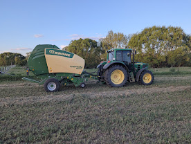 Green tractor pulling a green-and-yellow Krone round baler across a harvested field under a clear blue sky.