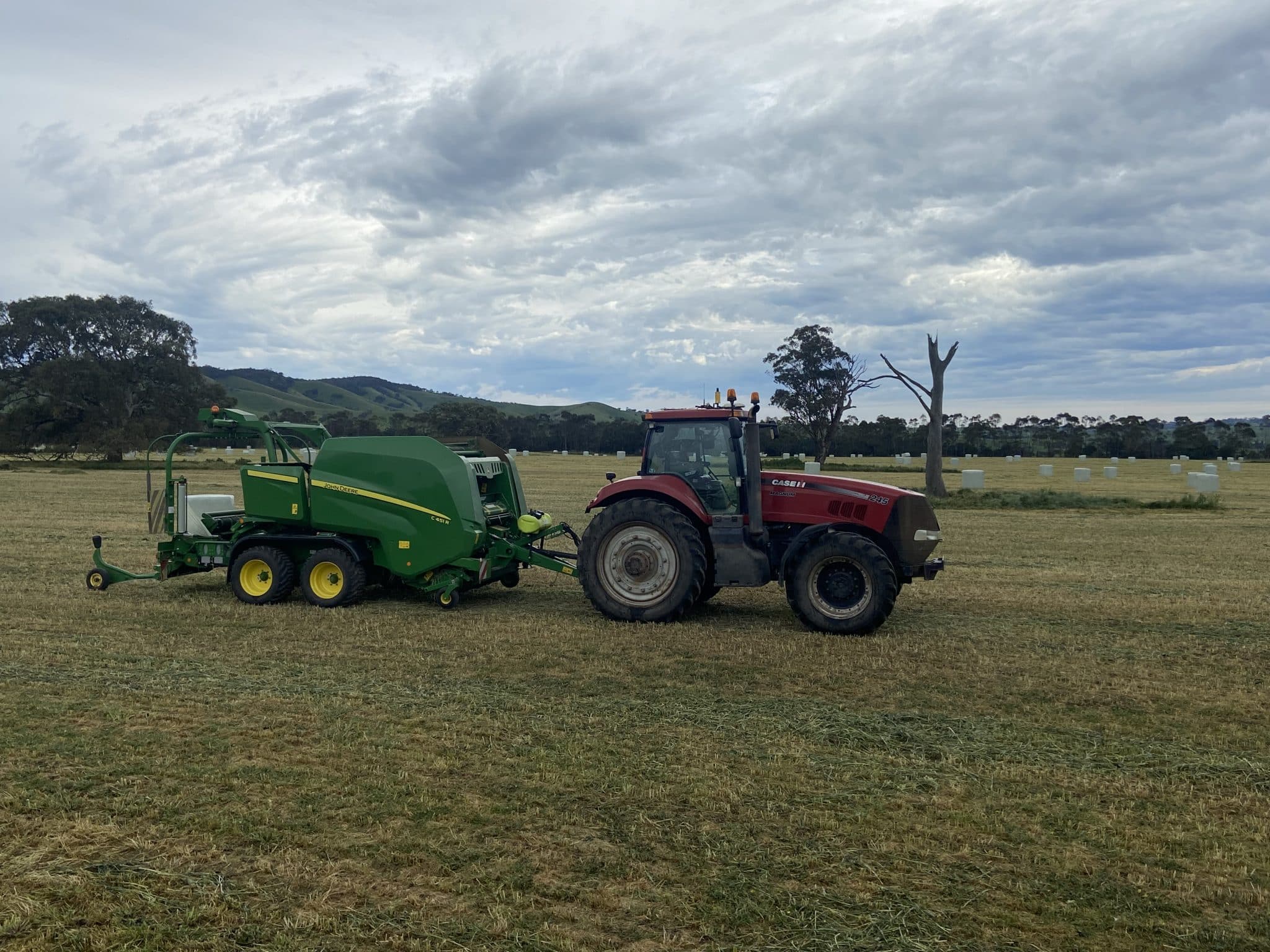Green John Deere baler attached to a red tractor sits in a harvested field under a cloudy sky, with distant hay bales and trees on the horizon.