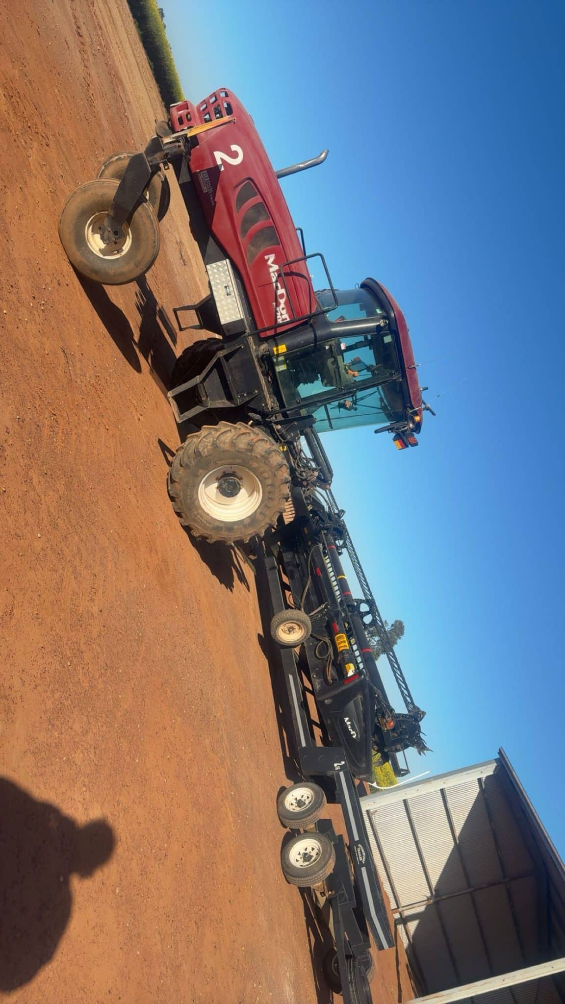 Red combine harvester on a dusty field with a clear blue sky, large tires and a cab visible beside a trailer area.