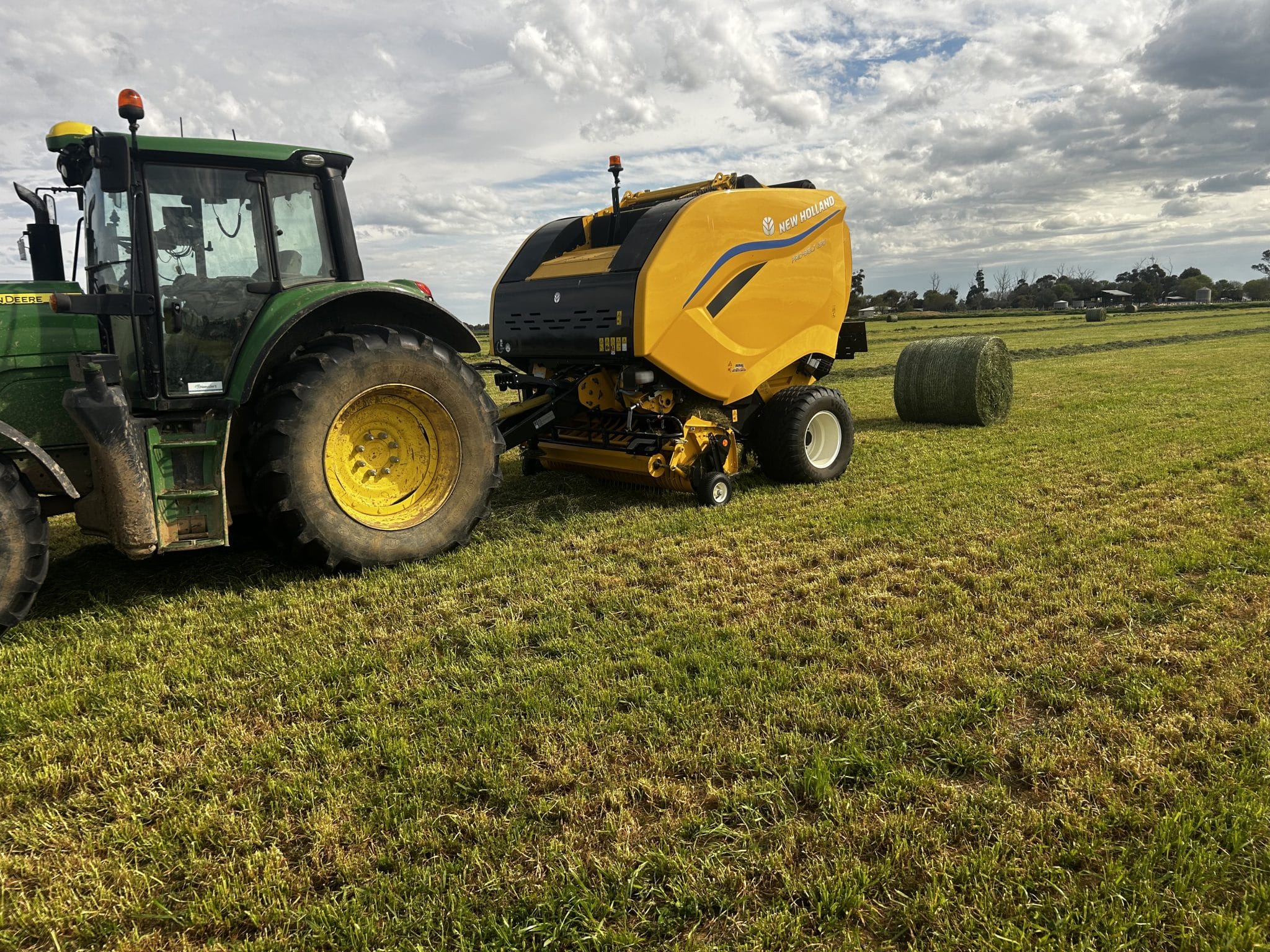 Green John Deere tractor hitched to a bright yellow New Holland baler in a grassy field with round hay bales in the distance.