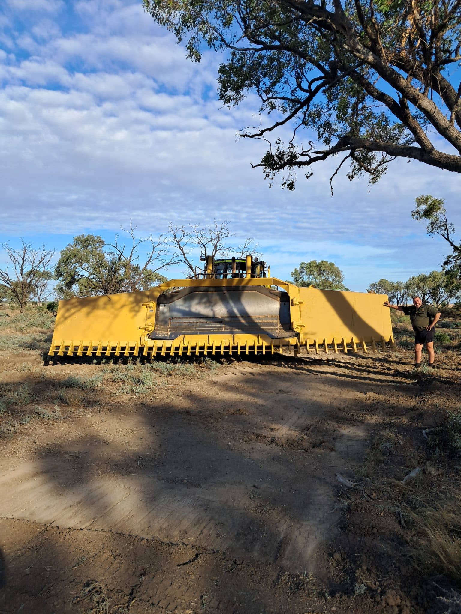 A large yellow industrial machine (bulldozer) in a dry, open landscape with a person leaning on the right side of it under a blue sky with scattered clouds.
