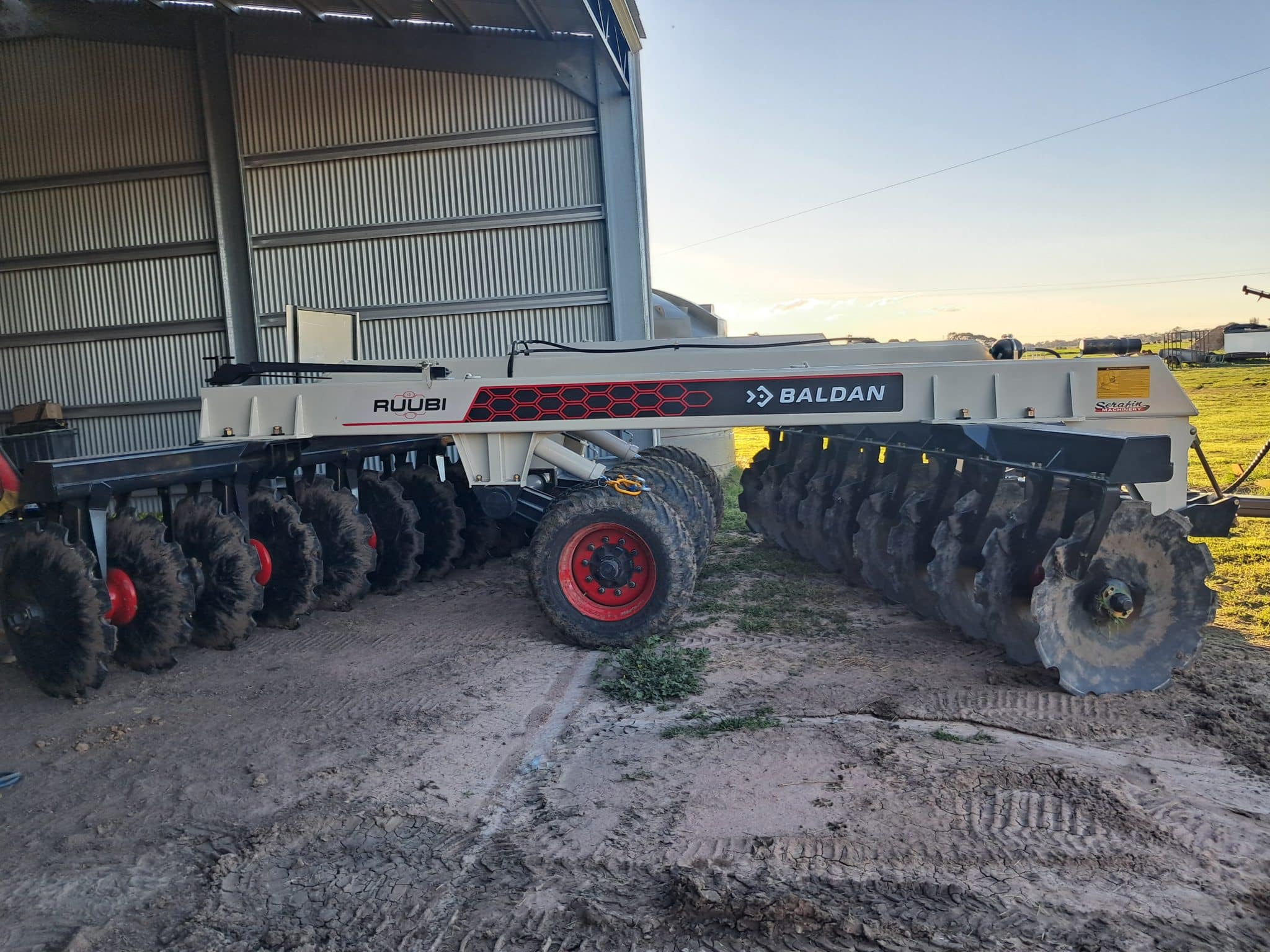 Large agricultural planter/seeder with red wheels parked beside a metal shed on a dirt farm lot