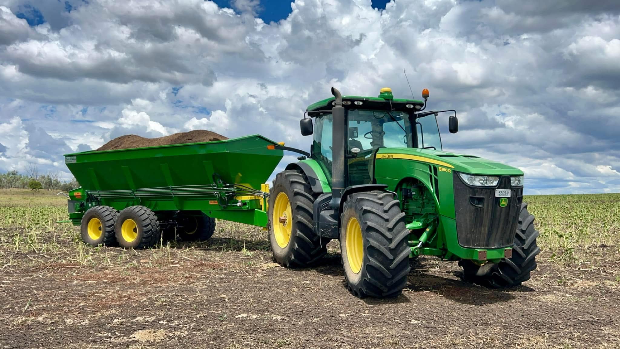 John Deere tractor with a large green grain cart attached in a plowed field under a cloudy sky