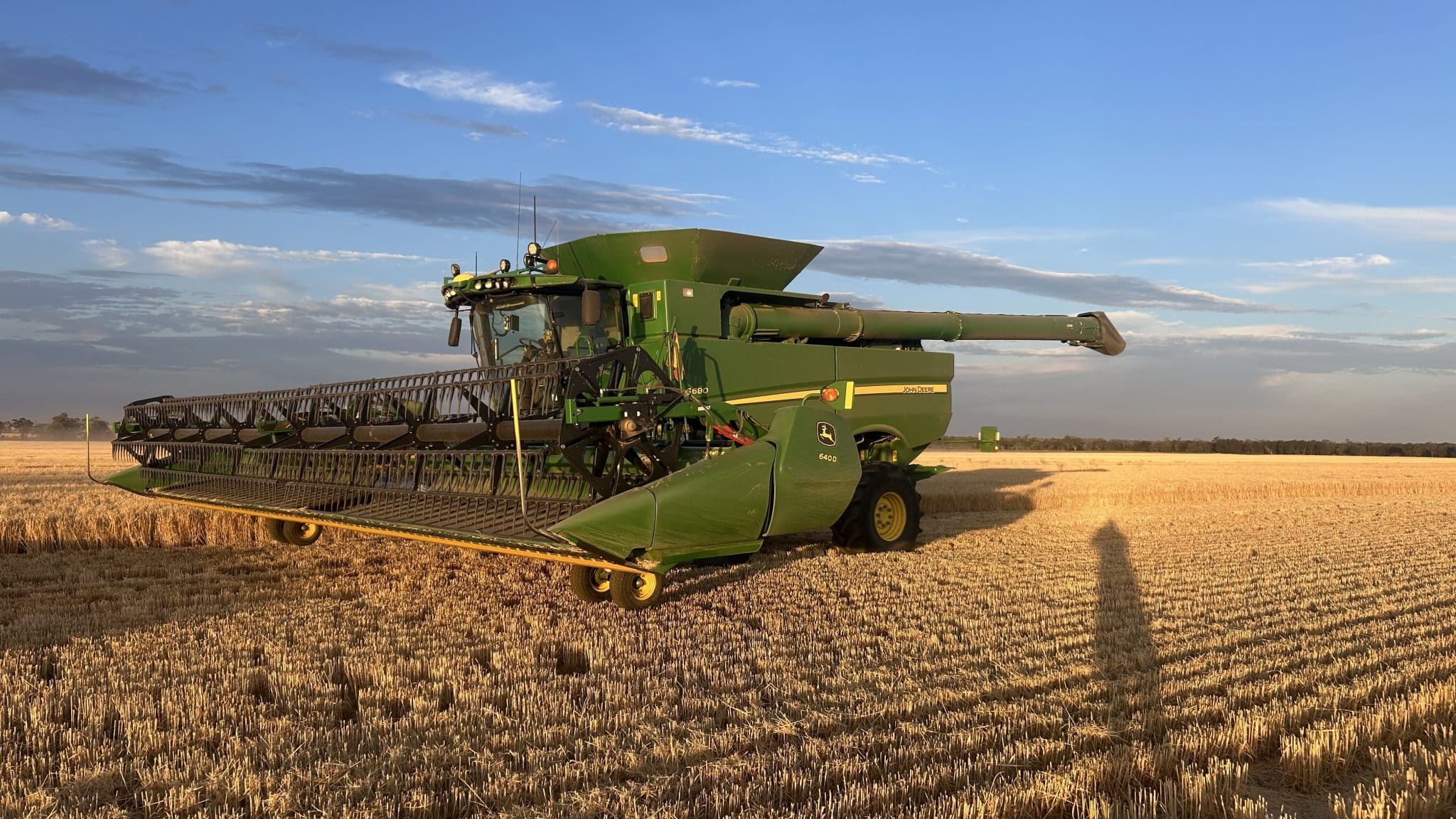 Green John Deere combine harvester in a harvested field under a blue sky during golden hour; long cutting header extended across the foreground.