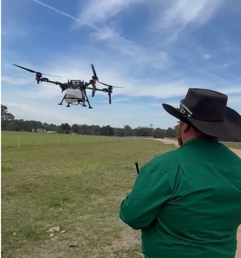 Person in a green shirt and wide-brimmed hat controls an octocopter drone hovering over a grassy field.