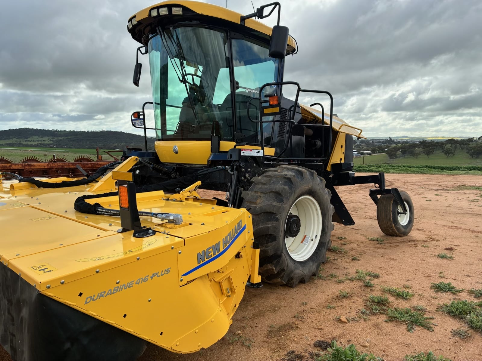 Yellow New Holland combine harvester on a dirt field with cloudy sky in the background, showing the front header and large tires.