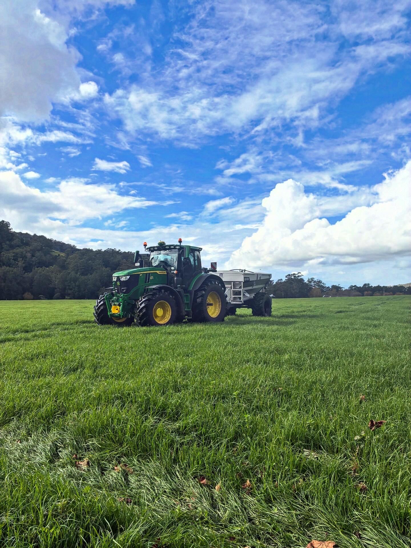 Green John Deere tractor with a silver trailer parked in a sunlit grassy field under a bright blue sky with scattered clouds.