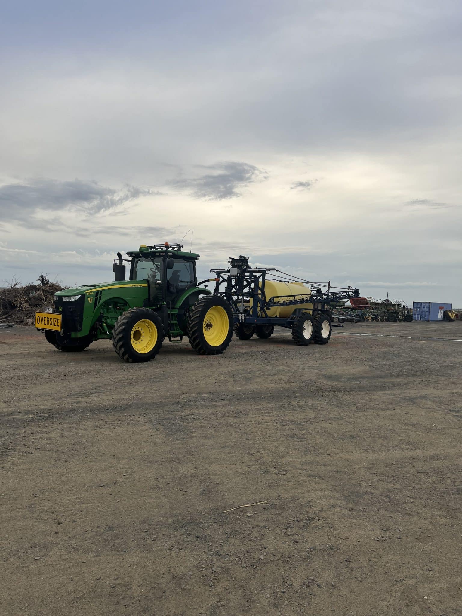 Green John Deere tractor with yellow wheels attached to farming equipment in a dirt lot under a cloudy sky (overcast day)
