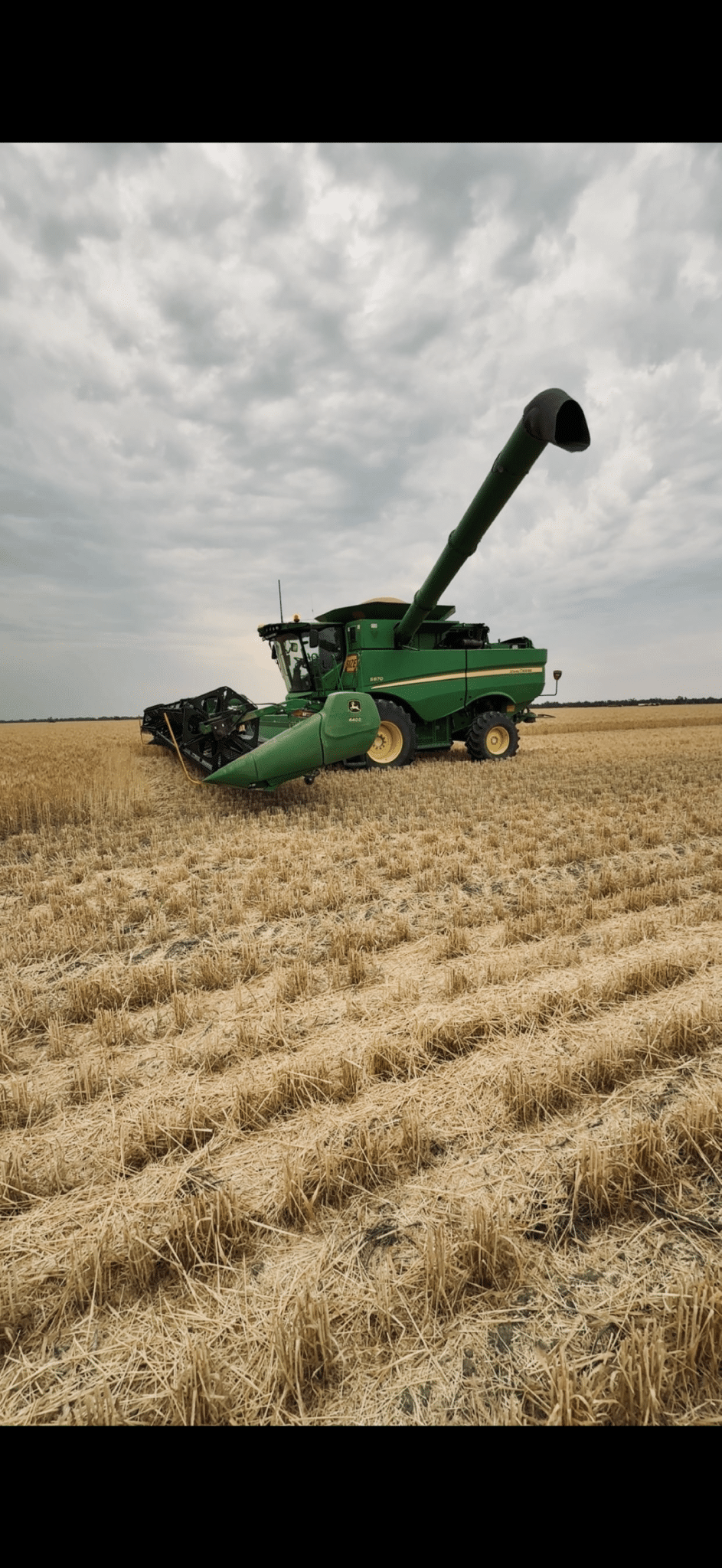 Green John Deere combine harvester in a harvested wheat field under a cloudy sky