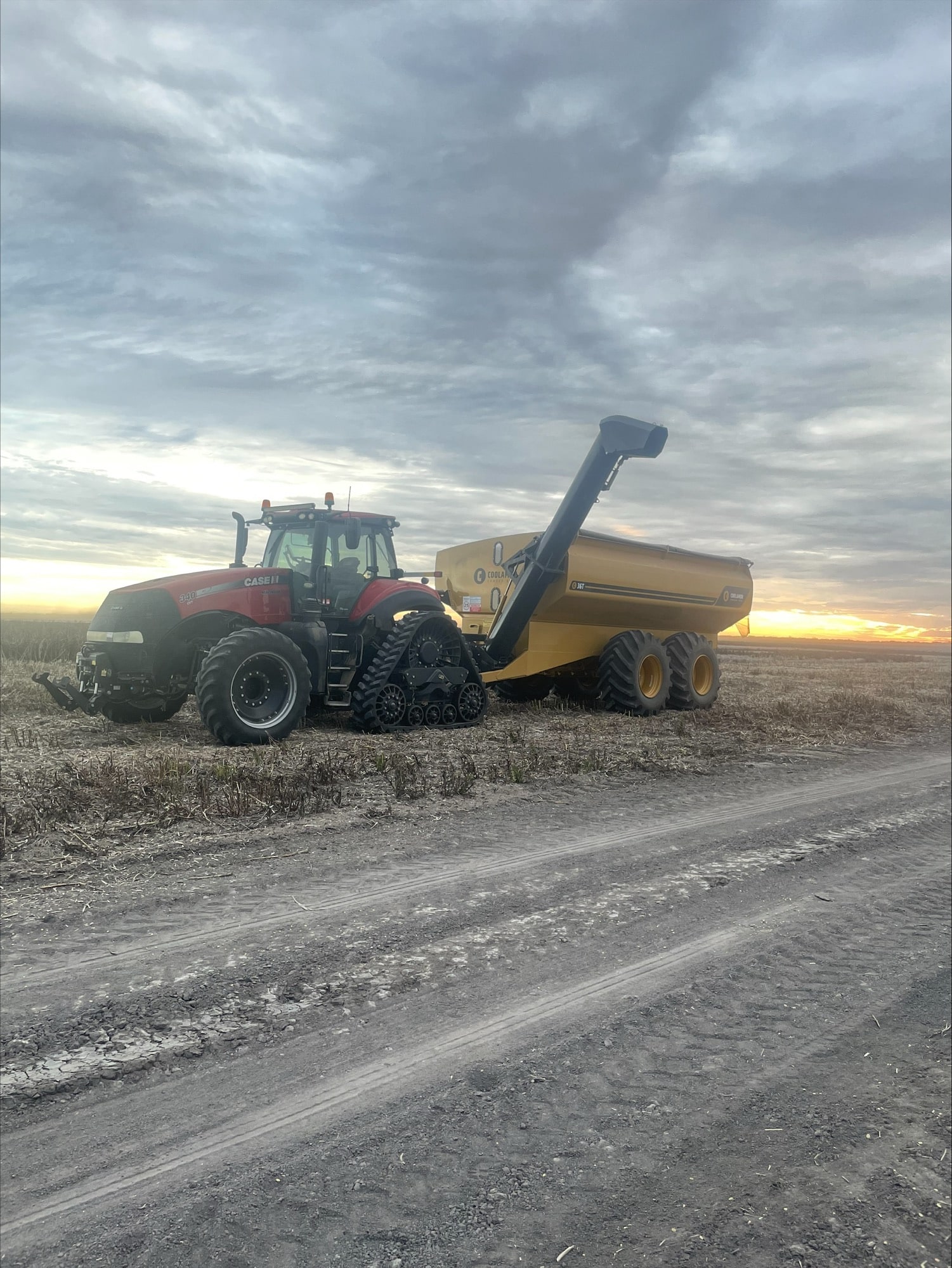 Red tractor connected to a yellow grain cart on a dusty farm field at sunset behind a cloudy sky.