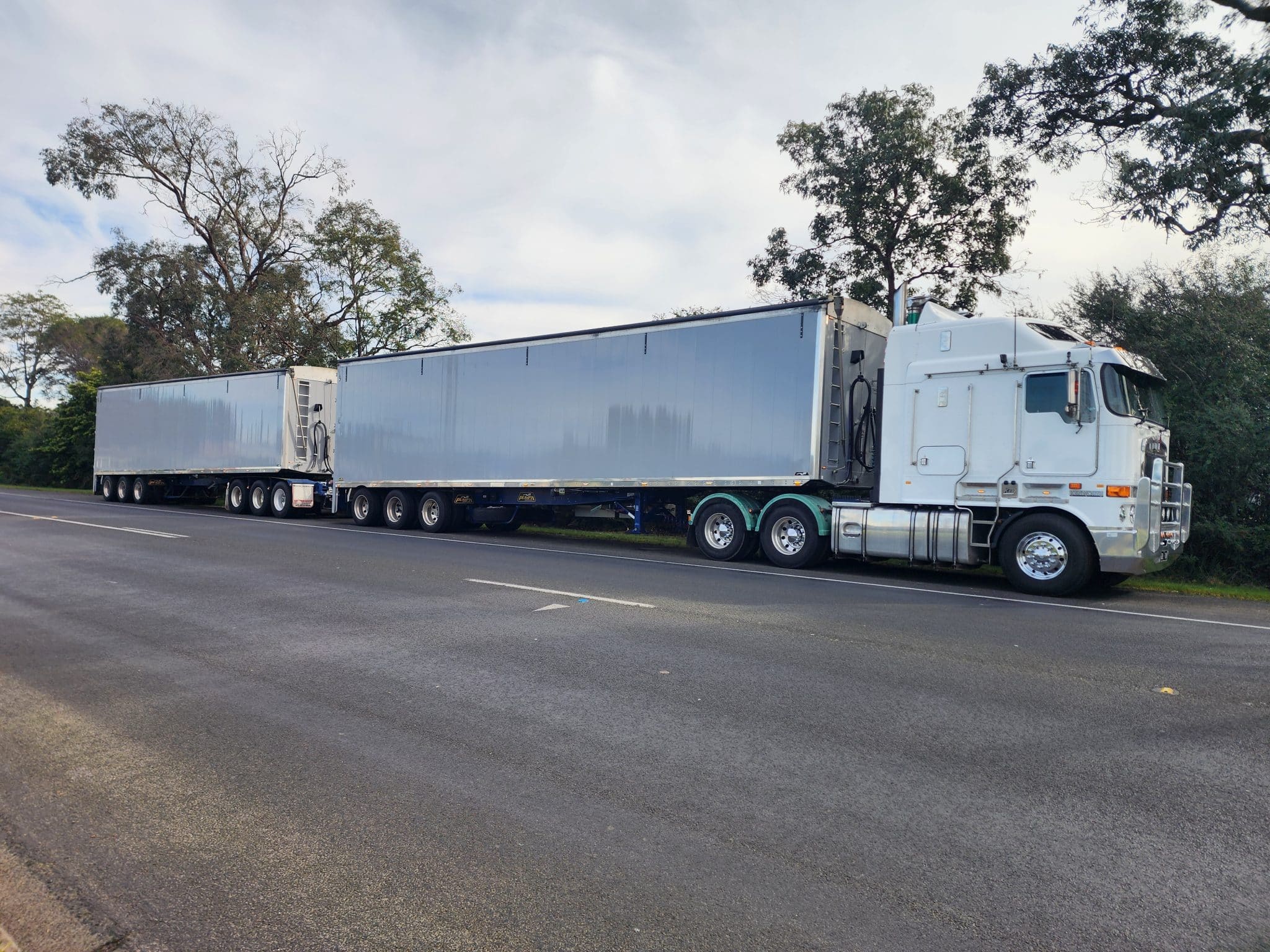 White semi-truck with two long silver trailers parked along a tree-lined road, under a cloudy sky.