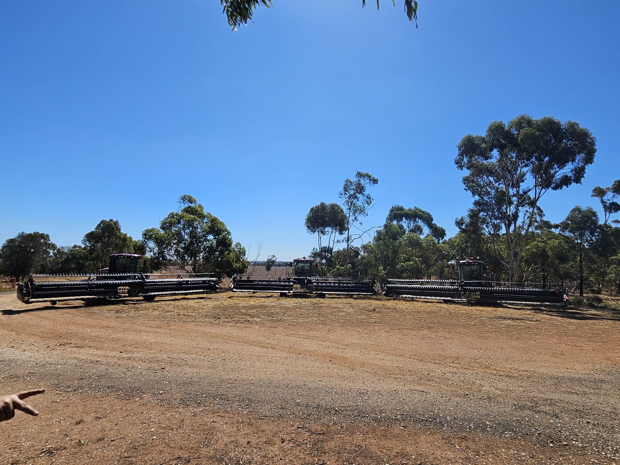 Wide view of a dirt field with stacked farming machinery parked near trees under a clear blue sky on a sunny day.