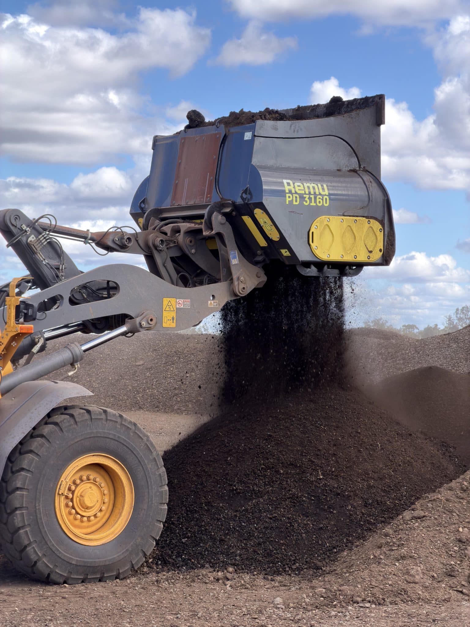 Remu PD 3160 screening bucket dumping dark gravel onto a pile at a quarry under a blue sky with clouds