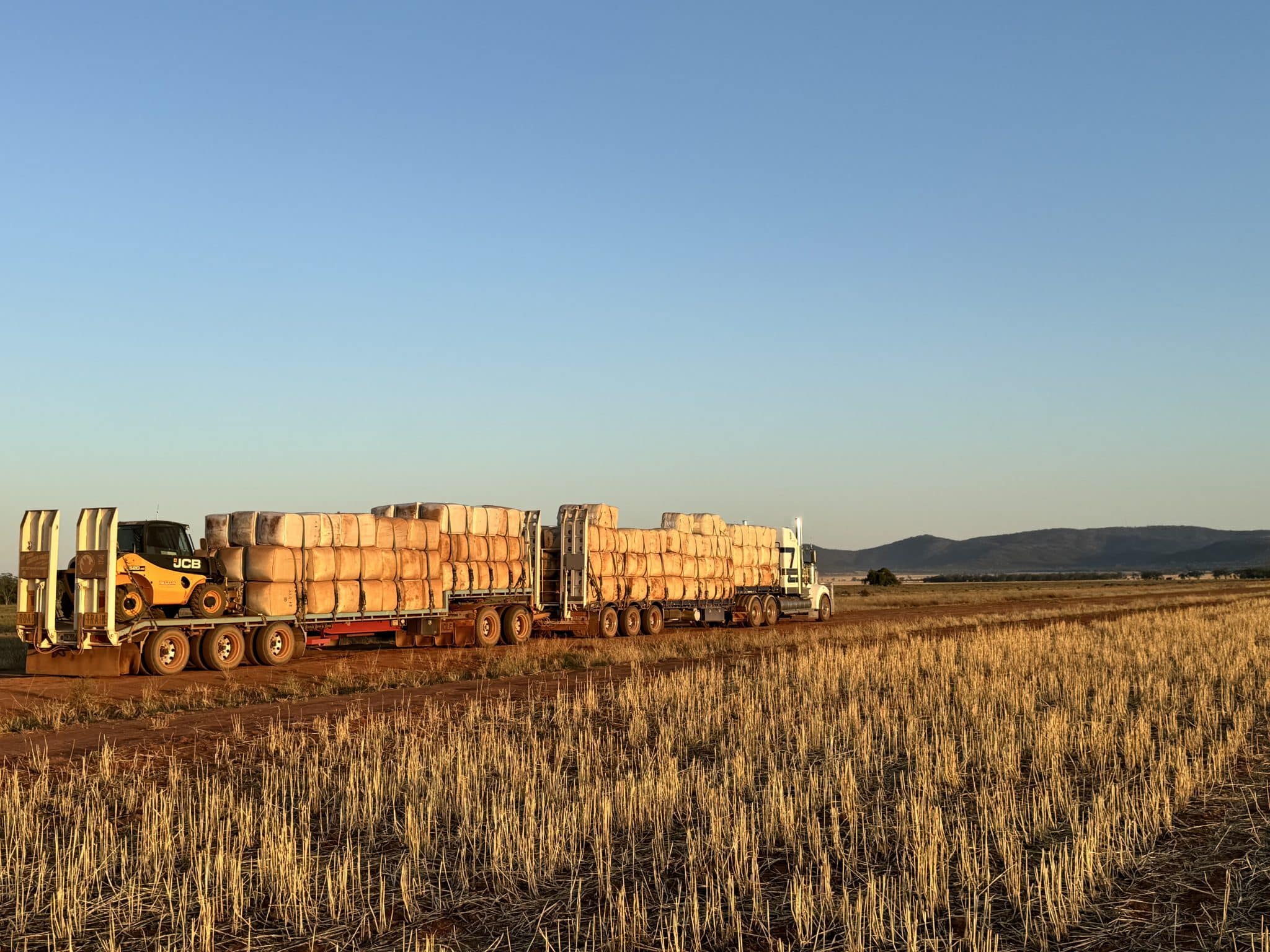 Long flatbed trailer carrying neatly stacked hay bales across a rural field at sunset under a clear sky