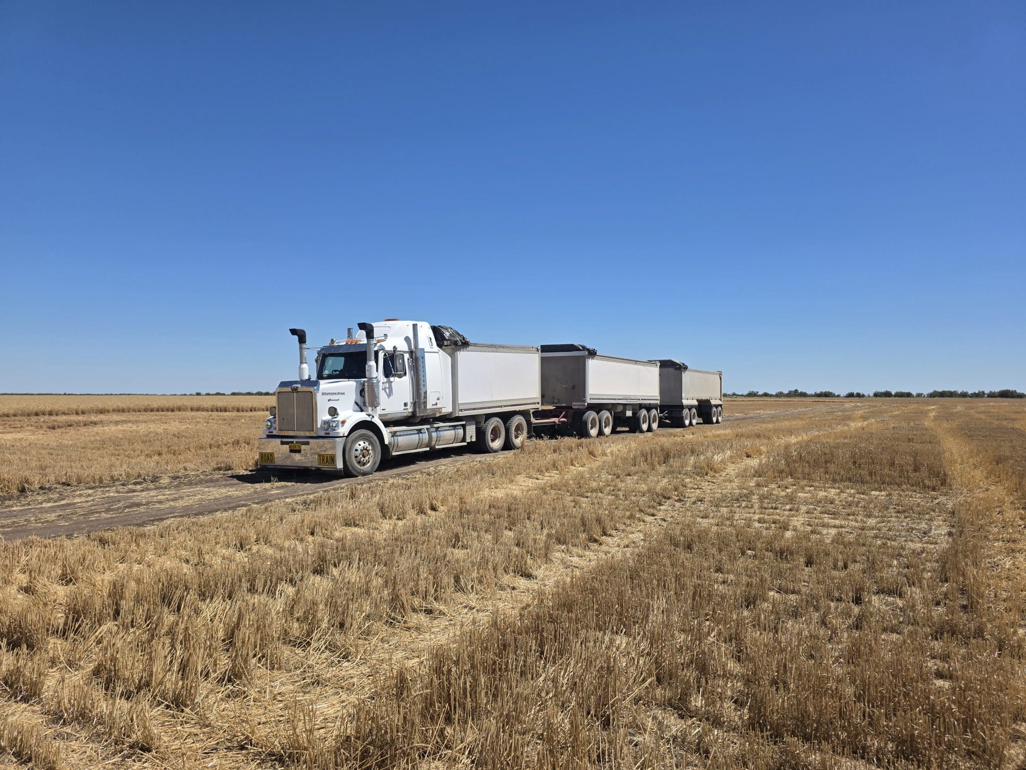 White semi-truck pulling two trailers on a dusty rural road under a clear blue sky in a harvested field.