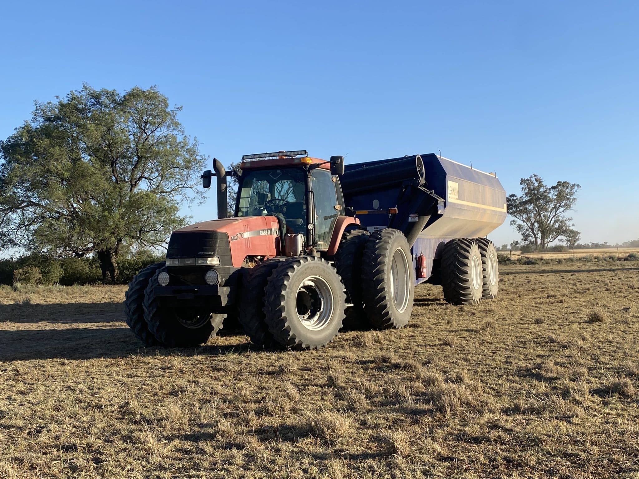 Red tractor with a blue dump trailer parked on a dry farmland under a clear blue sky, with trees in the background.