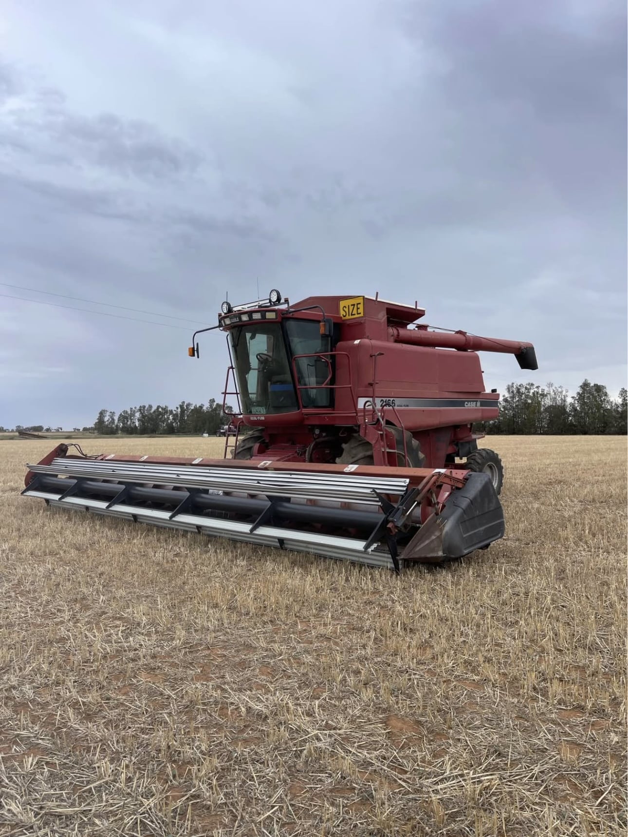 Red combine harvester with a wide cutting header parked in a harvested field under a cloudy sky, ready for use.