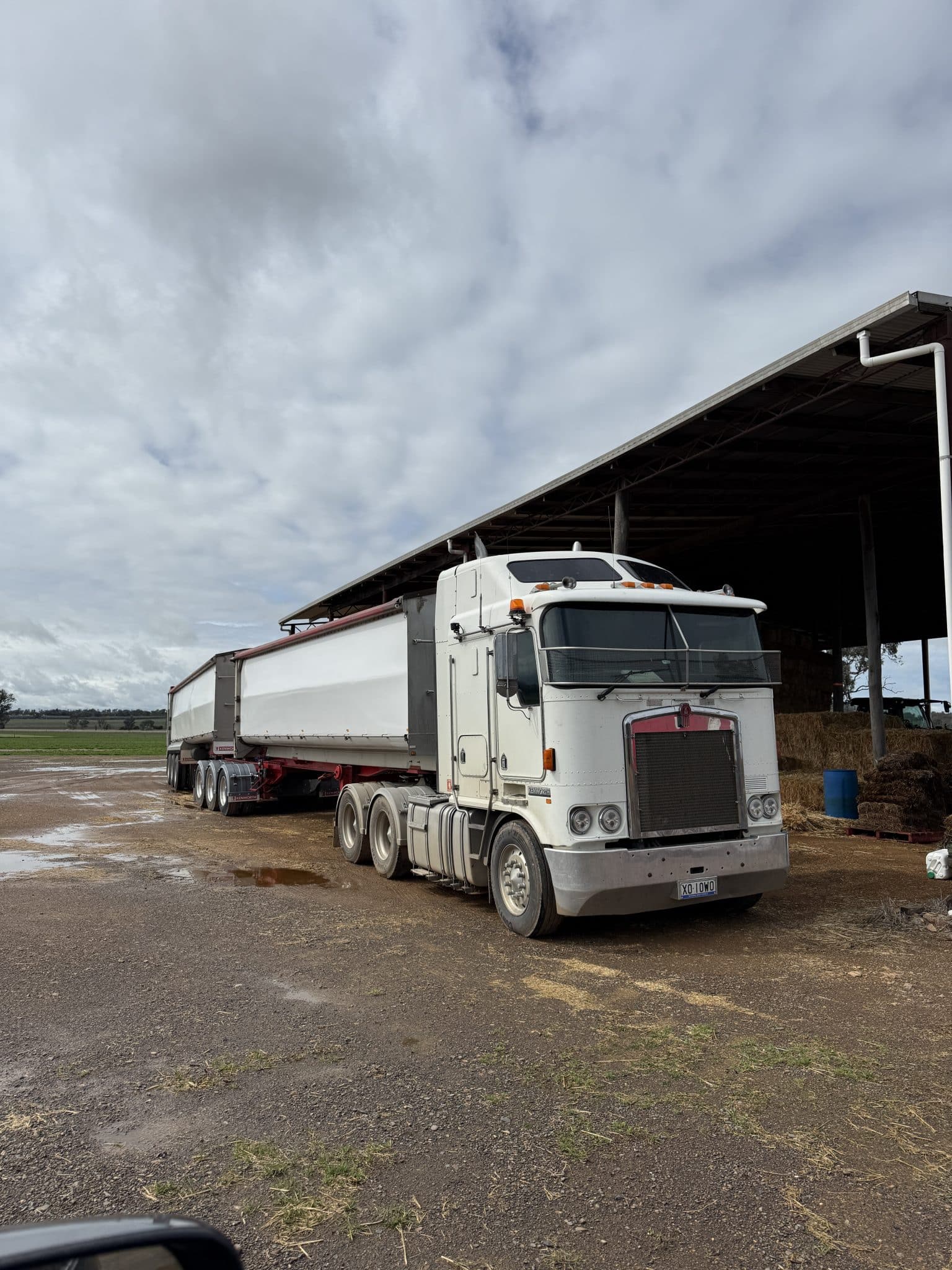 White semi-truck with a long trailer parked beside a barn under a cloudy sky on a muddy lots.