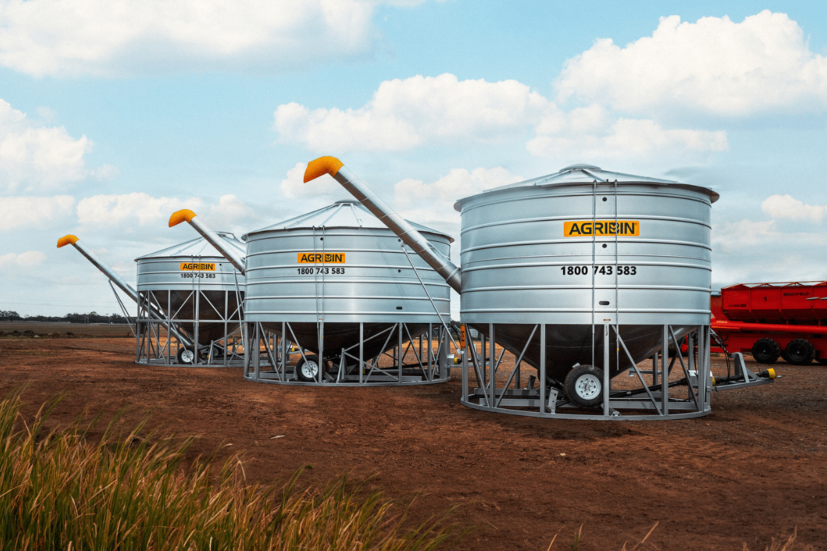 Row of large silver grain silos on metal frames with orange augers extending from their tops under a blue sky and clouds