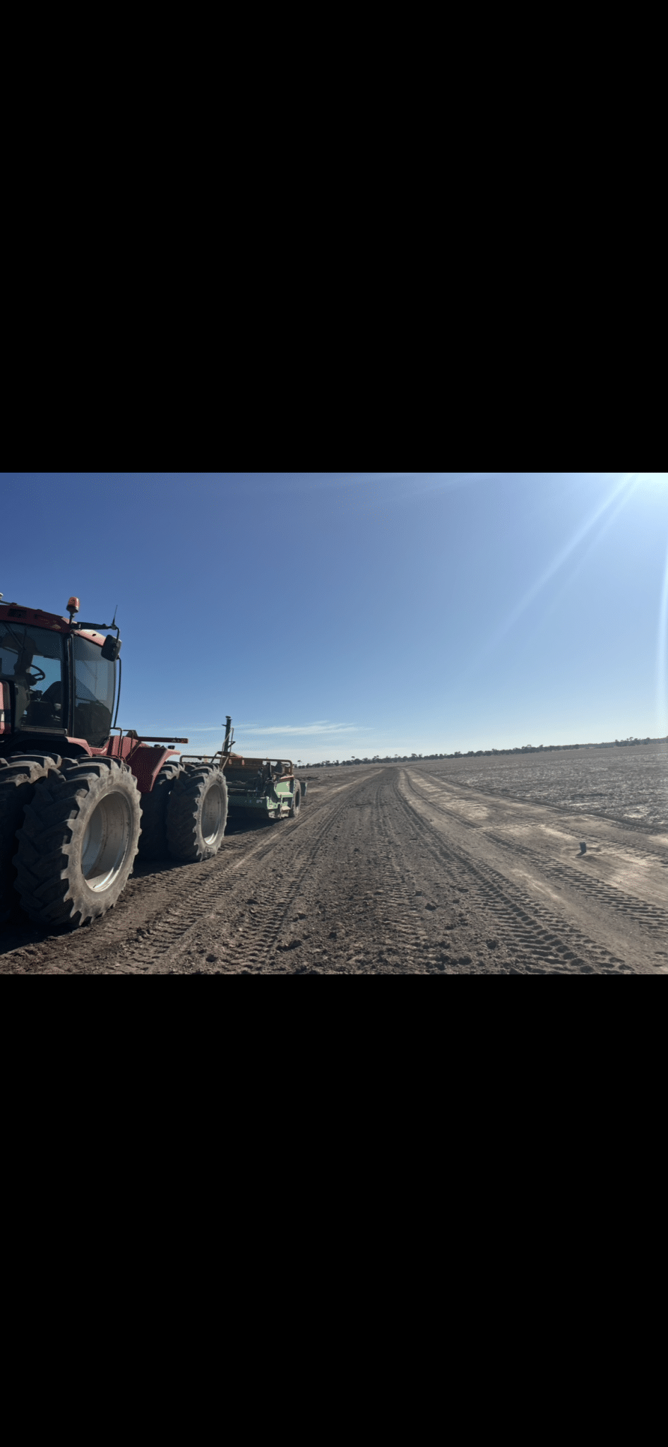 Red farm tractor with large tires sits at the edge of a freshly tilled field under a bright blue sky, sun glare to the right.