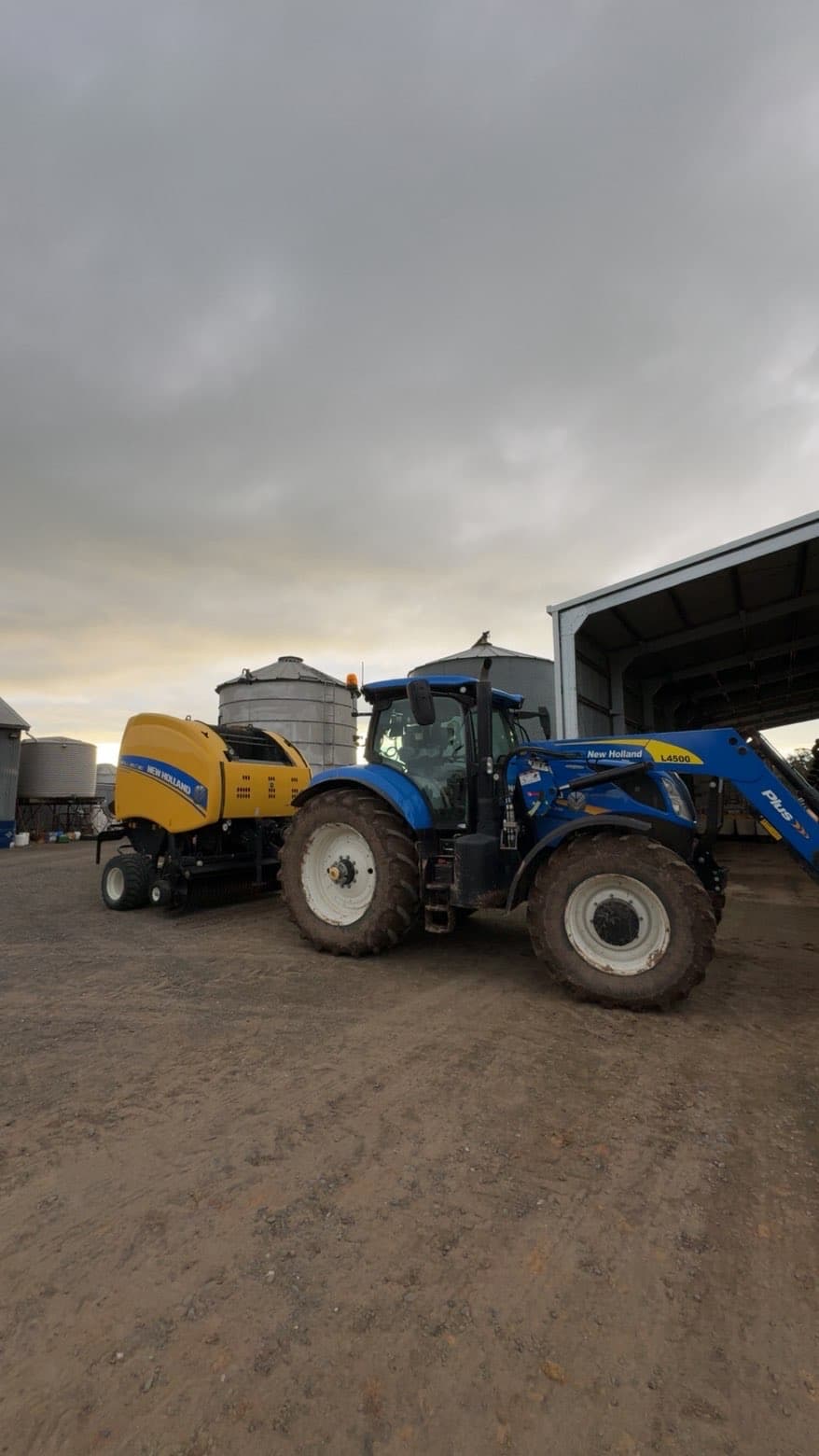 Blue New Holland tractor with front loader parked beside a yellow baler on a farm yard near grain silos in the background.
