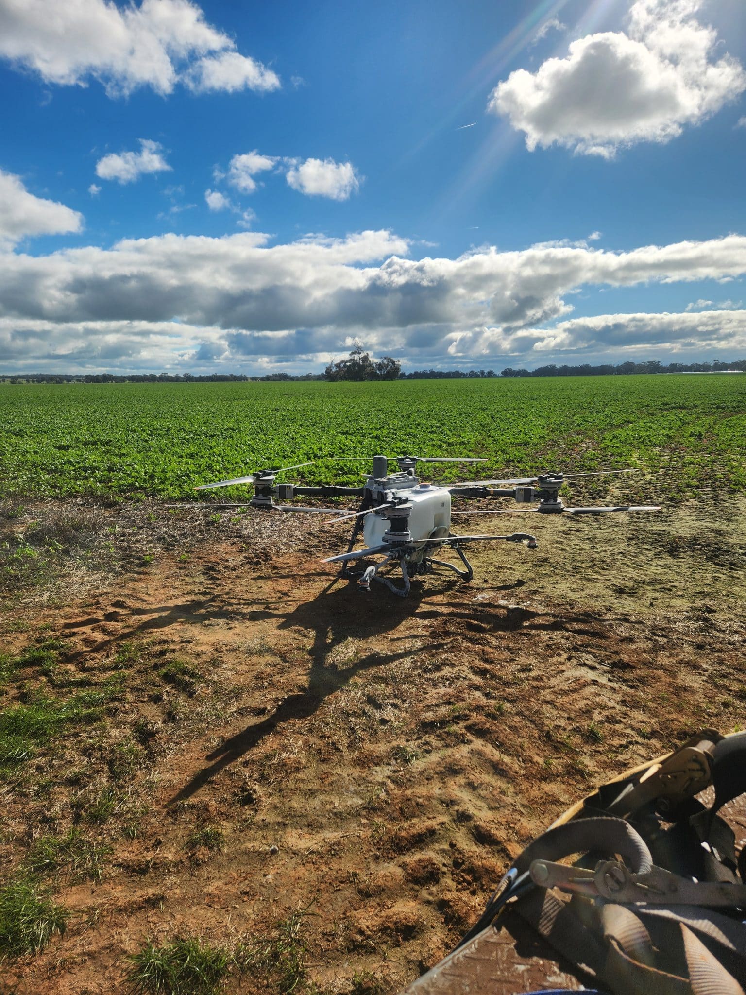 A drone sits on a dirt path at the edge of a green crop field under a blue sky with scattered clouds.