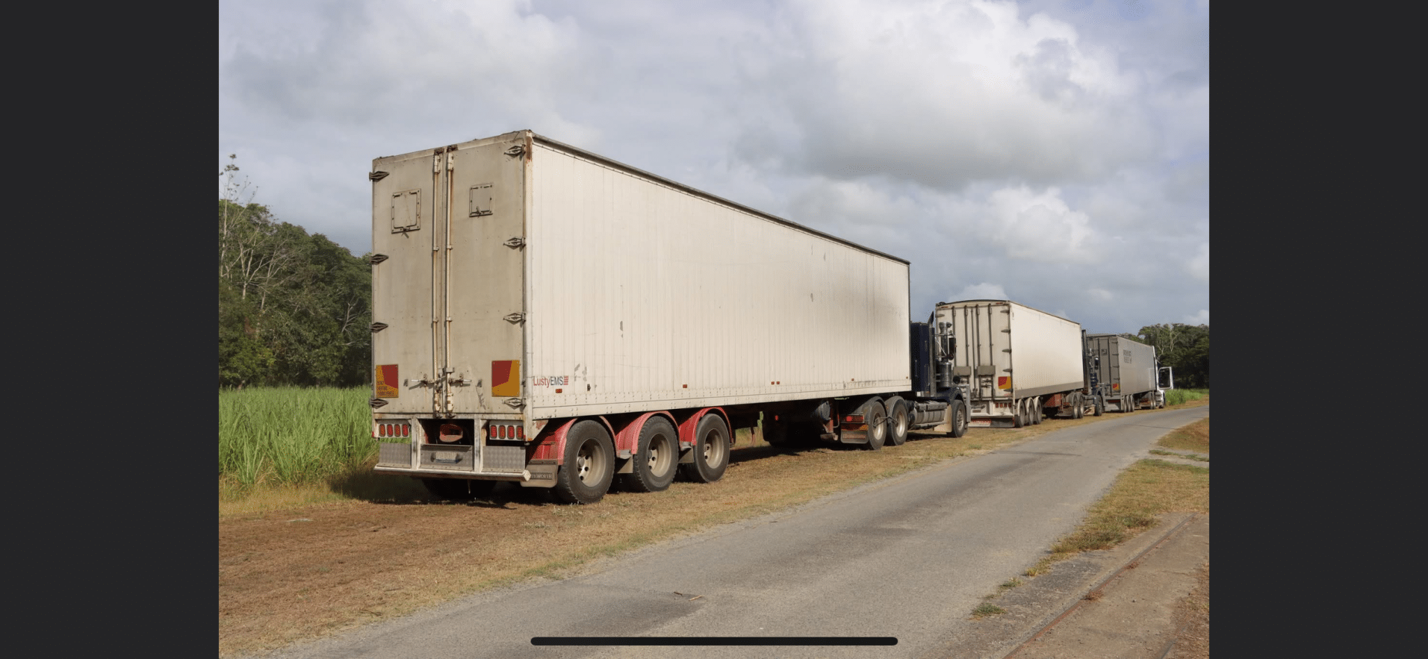 Line of large white semi-trailers parked along a rural road with fields and trees to the left and a cloudy sky above.
