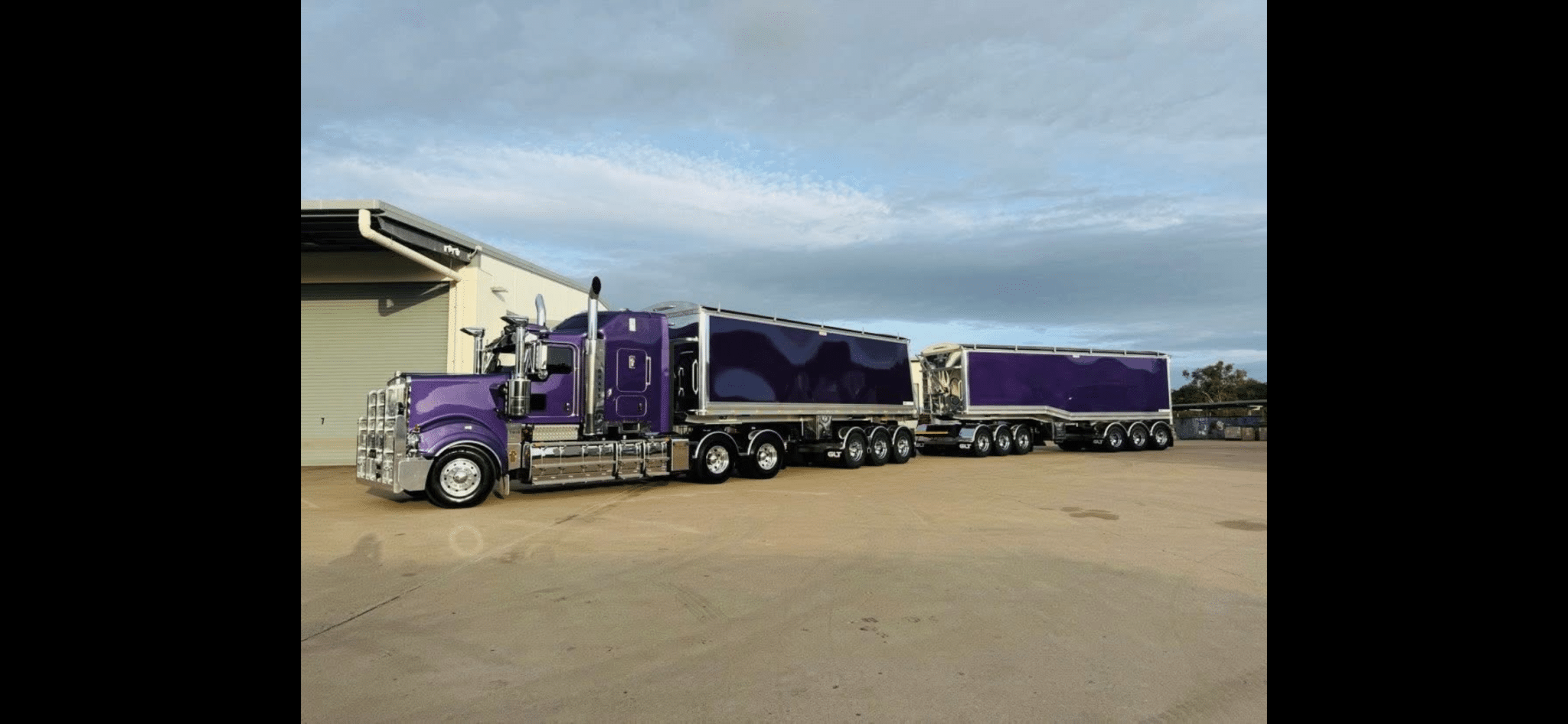 Purple semi truck with chrome accents and two matching purple trailers parked beside a beige warehouse in an industrial lot.