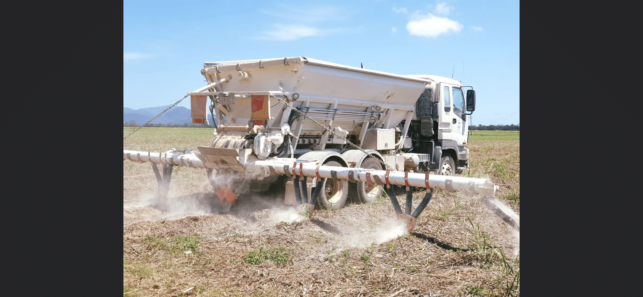 White agricultural sprayer truck in a dry field, extending a long boom to spray crops nearby.