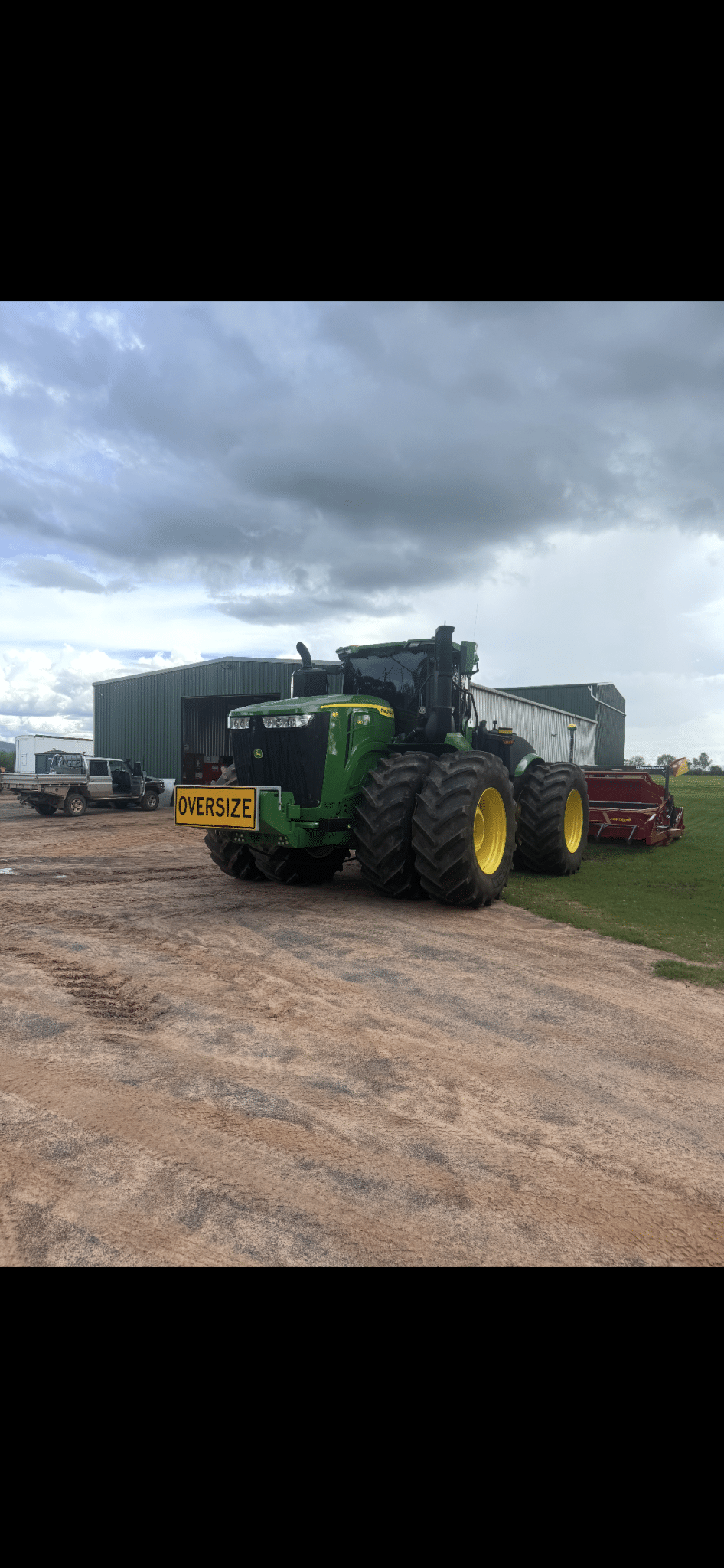 Large green John Deere tractor with an oversized load sign parked on a dirt yard near a barn, under cloudy skies.