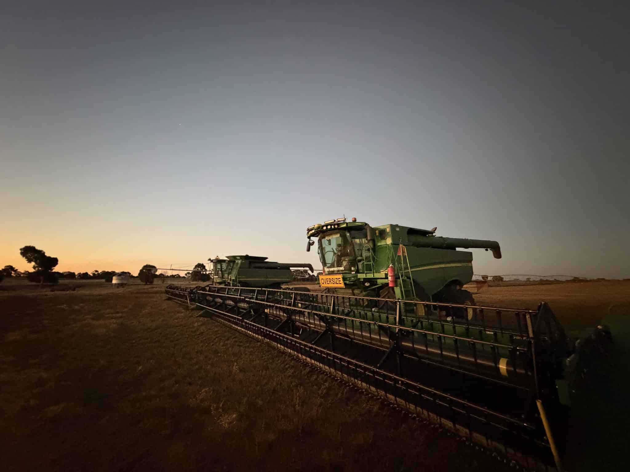Green combine harvester at dusk with an oversize sign and machinery on a farm field, low horizon.