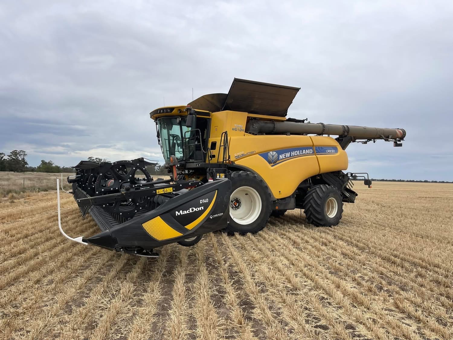 Yellow New Holland combine harvester in a harvested field, facing right with a MacDon header attached at the front